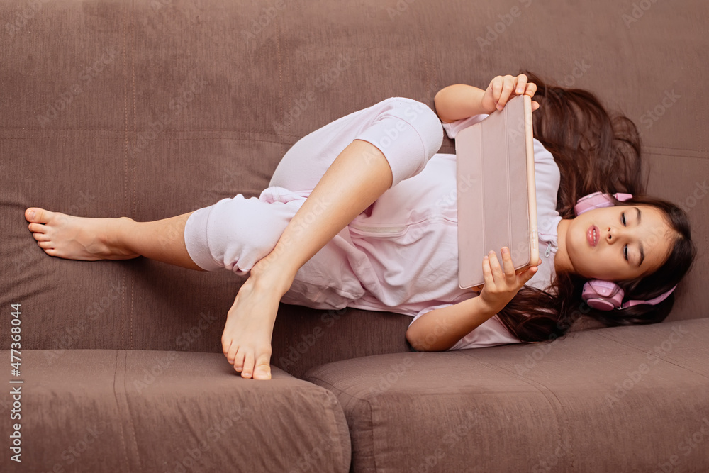 A little cute girl in pink home clothes and pink headphones, lies on the couch with digital tablet