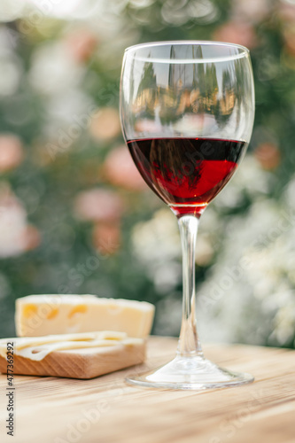 half-empty glass of red wine, piece of Maasdam cheese and slice of Maasdam cheese on bread on wooden brown table in sunny day at sunset in garden against organic background. selective focus. vertical