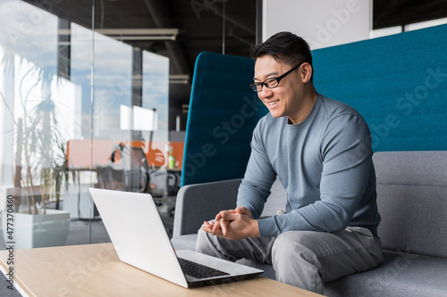 Asian with glasses, working in the office, making a video call with a laptop, smiling and rejoicing, communicating remotely with colleagues online, business man working in the office