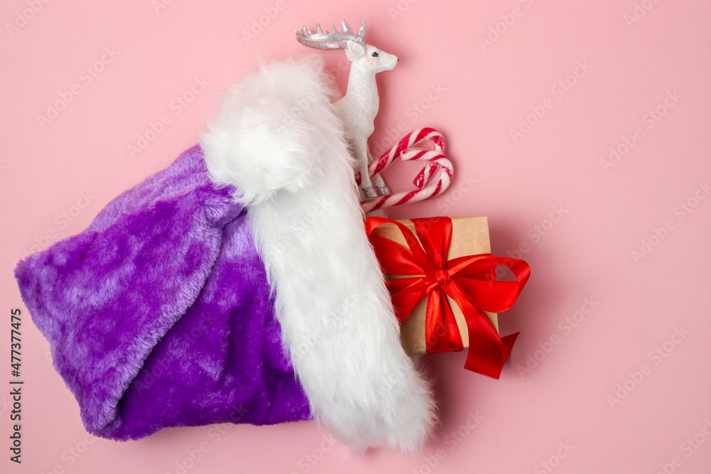 Christmas concept with candy canes, a box with a bow and a figurine of a white deer in a santa hat on a pink background. Flat lay, top view. Christmas concept with candy canes, a box with a bow and a figurine of a white deer in a santa hat on a pink background. Flat lay, top view.