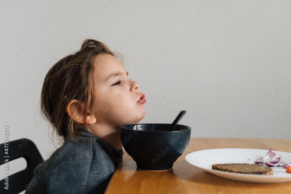 Unhappy child girl eats soup from black bowl with bread and onion. Lifestyle photo of kid in kitchen having a meal, Screaming kid. Picky eater Unhappy child girl eats soup from black bowl with bread and onion. Lifestyle photo of kid in kitchen having a meal, Screaming kid. Picky eater