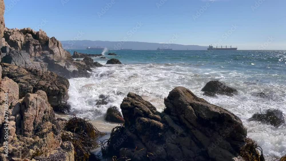 Waves crashing on rocks with cargo ships on sunny day in Valparaiso, Chile Waves crashing on rocks with cargo ships on sunny day in Valparaiso, Chile