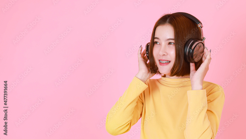 Portrait studio cutout shot of Asian young pretty short hair female model  in yellow long sleeve shirt standing holding adjusting big black headphones  while listening to music song on pink background Stock-Foto