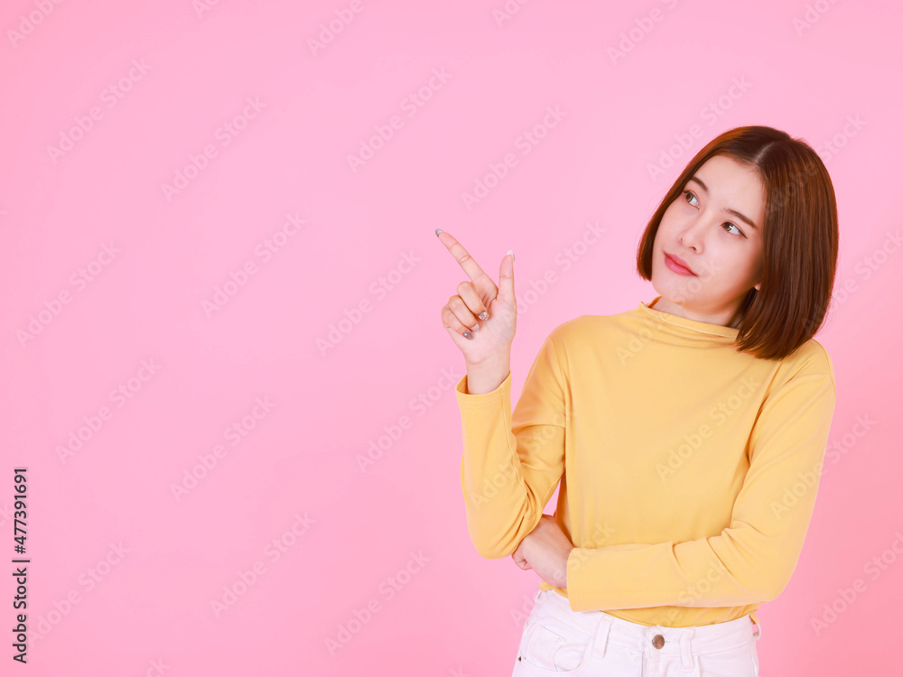 Portrait studio cutout shot of Asian young pretty short hair female model in yellow long sleeve shirt stand smile to look at the camera and pointing finger on pink background. Portrait studio cutout shot of Asian young pretty short hair female model in yellow long sleeve shirt stand smile to look at the camera and pointing finger on pink background.