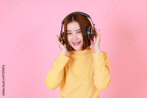 Portrait studio cutout shot of Asian young pretty short hair female model in yellow long sleeve shirt standing holding adjusting big black headphones while listening to music song on pink background