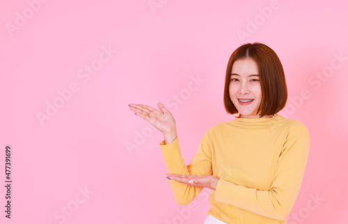 Portrait studio cutout shot of Asian young pretty short hair female model in yellow long sleeve shirt stand smile to look at the camera and pointing finger on pink background.
