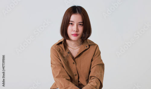 Studio cutout shot of Asian thoughtful young pretty short hair female model in long brown coat jacket casual shoes sitting crossed legs thinking decide solve idea on box chair on white background © Bangkok Click Studio