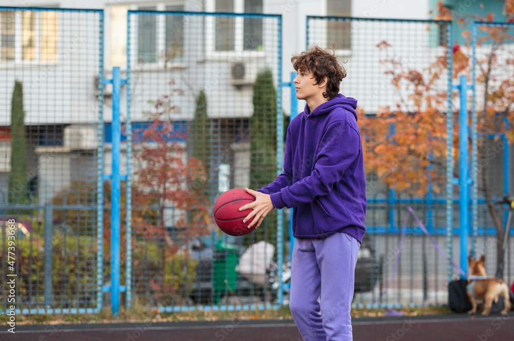 Cute teenager in violet hoodie playing basketball. Young boy with ball learning dribble and shooting on the city court. Hobby for kids, active lifestyle Cute teenager in violet hoodie playing basketball. Young boy with ball learning dribble and shooting on the city court. Hobby for kids, active lifestyle