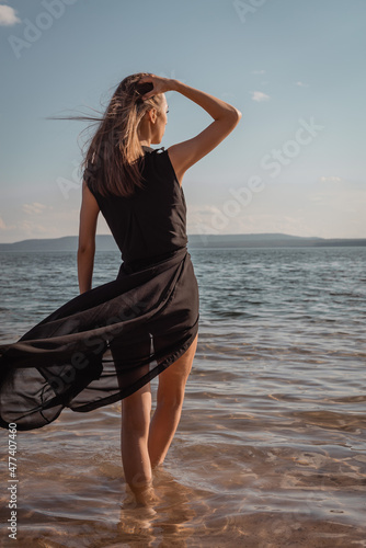 A young woman in a black dress stands in the water by the sea. High quality photo