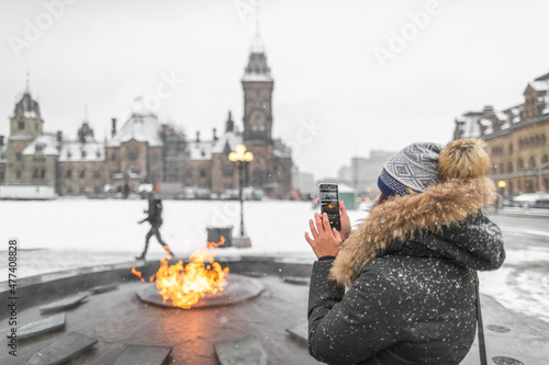 Ottawa travel tourist woman taking photo with phone of Canadian Parliament in Ontario, Canada. Snowing landscape and centennial flame that do not freeze in winter. photo