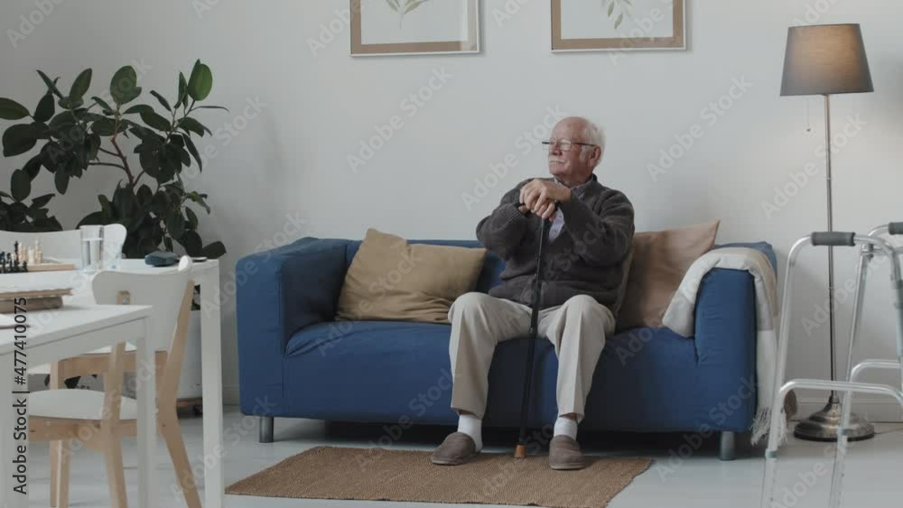 Full-length shot of senior Caucasian man with grey hair and moustache sitting on sofa with walking stick looking away Full-length shot of senior Caucasian man with grey hair and moustache sitting on sofa with walking stick looking away