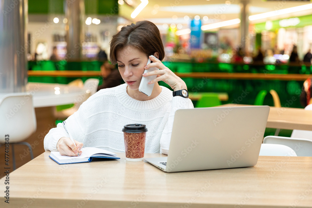 Young beautiful pretty brunette woman wearing white sweatshirt sitting at a shopping center at a table and working at a computer laptop, using mobile phone, writing information at notebook. Freelance