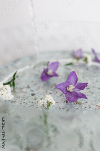 into a glass basin with purple flowers floating in the water. against the background of a ceramic table and a white wall. The concept of spa and self-care.