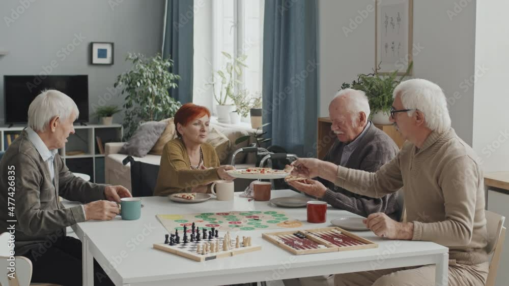 Medium shot of cheerful group of senior people sitting around table in nursing home eating pizza, talking and playing board games Medium shot of cheerful group of senior people sitting around table in nursing home eating pizza, talking and playing board games