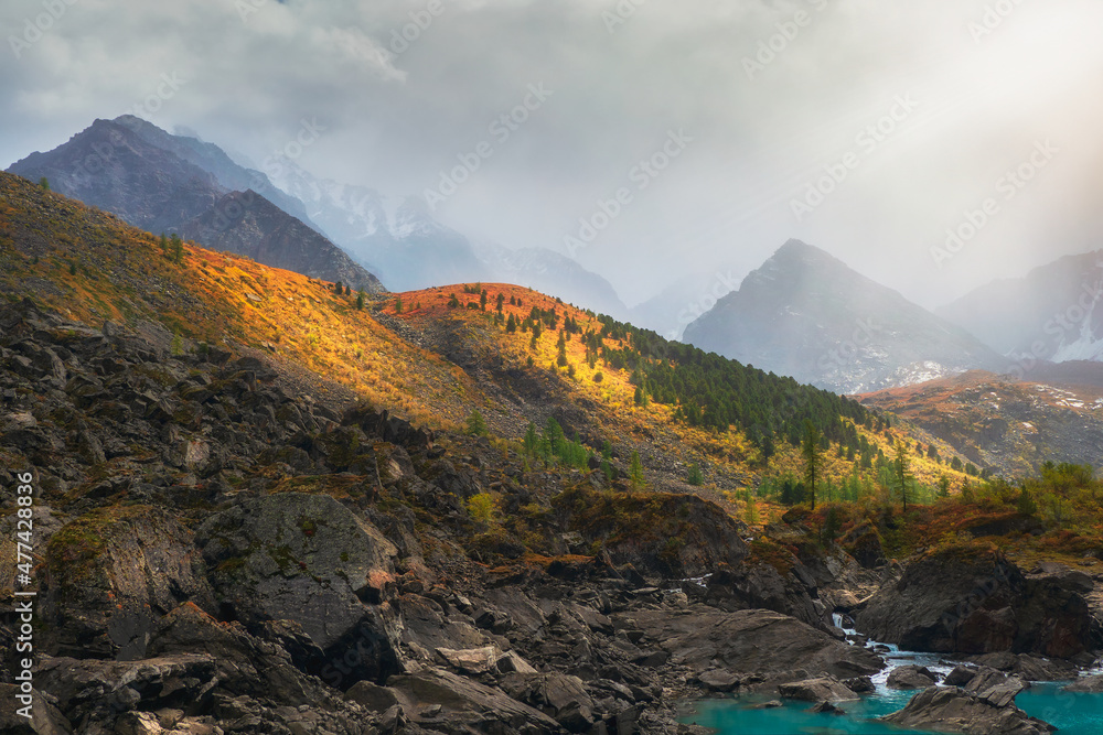Colorful mountain landscape with great mountain lit by dawn sun among dark clouds. Awesome alpine scenery with high mountain pinnacle at sunset or at sunrise. Big mountain in orange light.