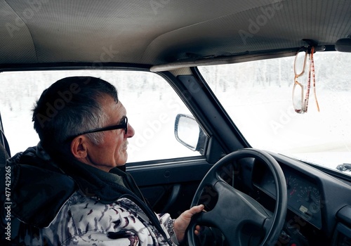 An elderly man in a winter jacket driving a Lada car goes fishing in winter. Winter and snowfall behind the glass.