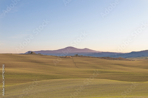 Photo of a Tuscan countryside panorama during sunset. In the foreground the cultivated fields and a wood, in the background some houses, the mountains and the sun's rays