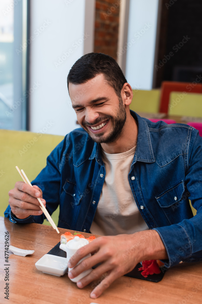 cheerful man laughing with closed eyes while holding chopsticks near sushi rolls cheerful man laughing with closed eyes while holding chopsticks near sushi rolls