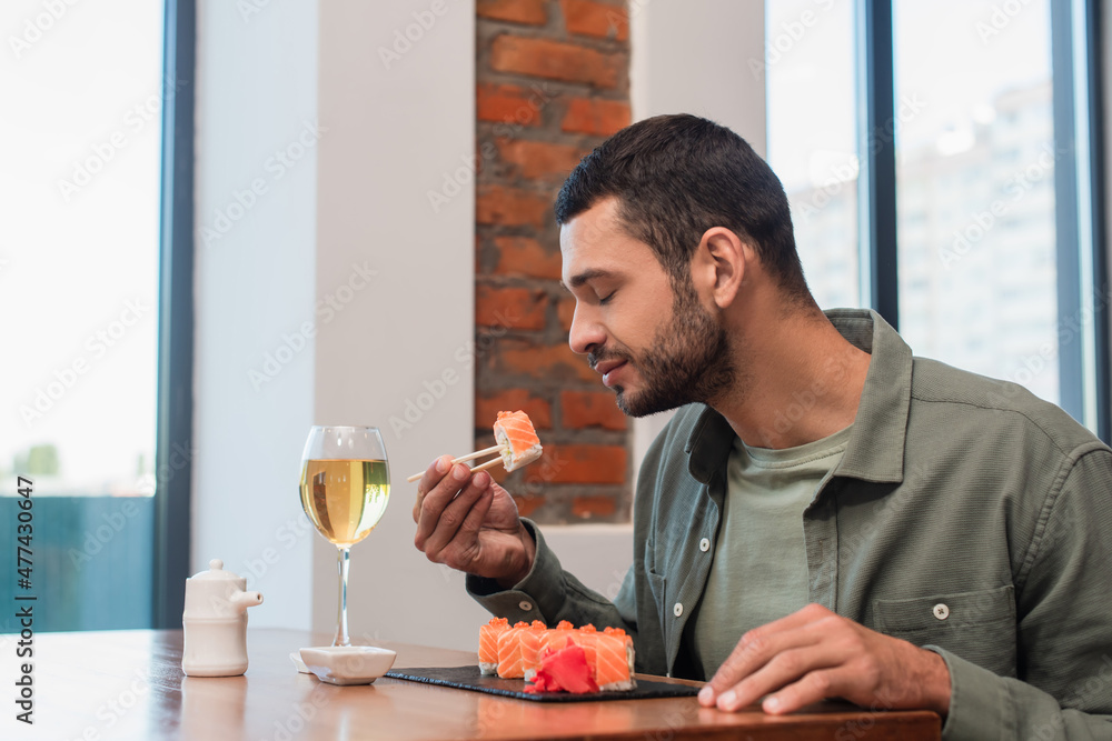 young man with closed eyes holding delicious sushi with chopsticks near glass or white wine young man with closed eyes holding delicious sushi with chopsticks near glass or white wine