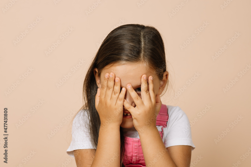 Little girl covering face with hands with slightly visible shining eyes and happy smile looking at camera wearing bright, pink jumpsuit and white t-shirt on beige background. Little girl covering face with hands with slightly visible shining eyes and happy smile looking at camera wearing bright, pink jumpsuit and white t-shirt on beige background.
