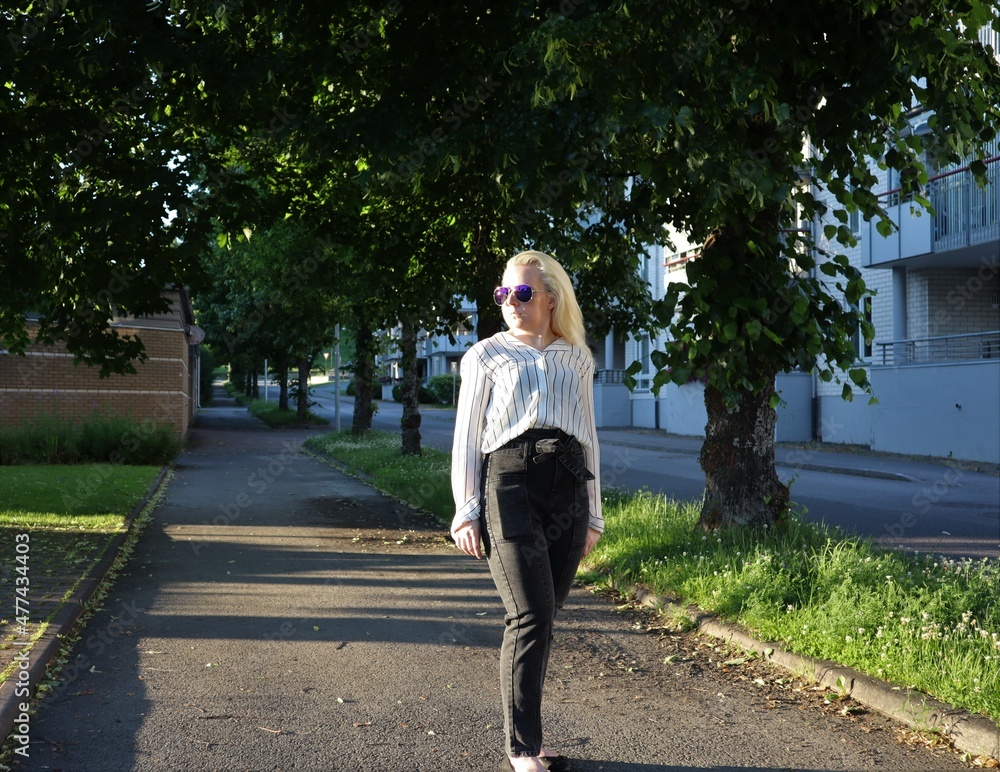 Adult woman portrait real estate architect agent with blue sunglasses in black and white stripes shirt and black high waist jeans pants taking a walk down the the neighborhood street during sunset