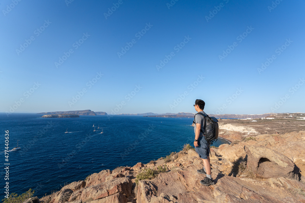 An Asian man standing on a cliff by the sea in Santorini