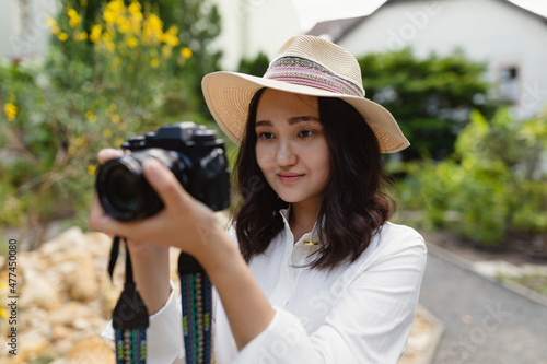 Young Asian woman in white shirt and hat in a park, smiling, holding camera. Hobby or professional photography concept. Spring or summer time. Outdoor portrait