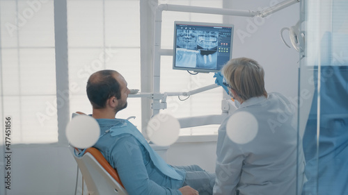 Mature dentist pointing at teeth radiography on monitor to explain dentititon diagnosis to patient with toothache. Dental expert showing x ray scan results to man with cavity pain.