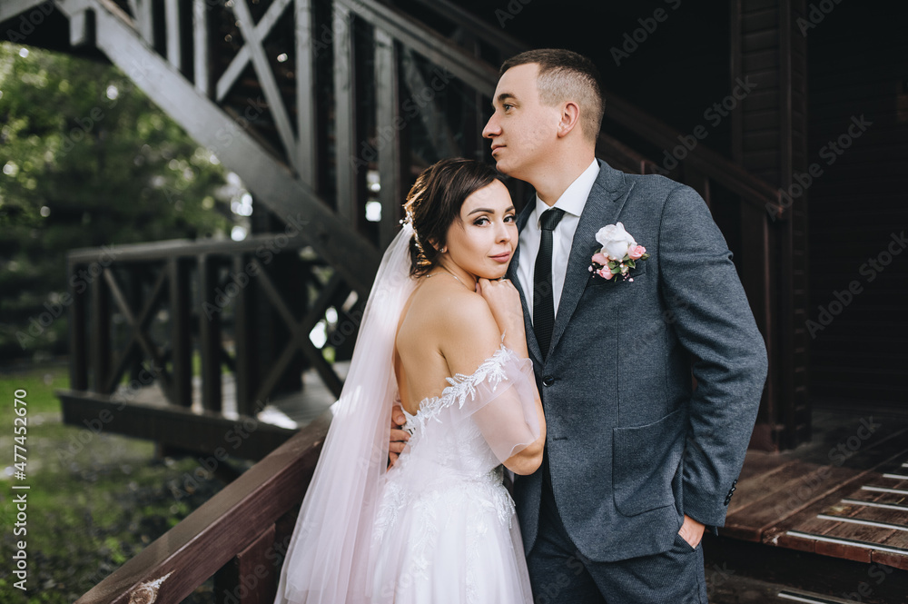 Stylish groom and a beautiful brunette bride in a white dress with a bouquet in their hands are standing and hugging on the background of a wooden house with a ladder. Wedding portrait. Stylish groom and a beautiful brunette bride in a white dress with a bouquet in their hands are standing and hugging on the background of a wooden house with a ladder. Wedding portrait.