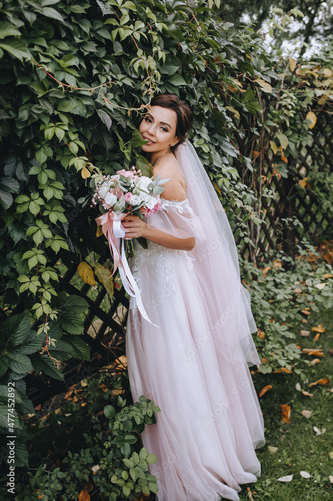 Wedding portrait of a beautiful, smiling brunette girl with a bouquet in her hands, standing on the grass, against the background of green ivy foliage of nature.