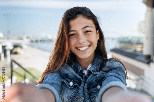 Cute, positive teenager holding a camera, looking straight. Beautiful girl sincerely smiling on the background of the city