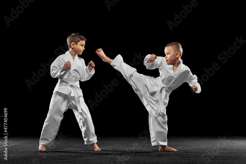 Two little kids, boys, taekwondo athletes training together isolated over dark background. Concept of sport, education, skills