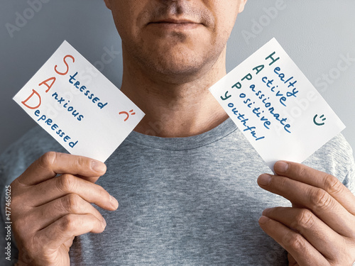 Middle aged man holding cards with Happy or Sad mood descriptions. Concept of choosing how person can spend this day depends on per sonal choice. Grey background and t-shirt. Focus on hands photo