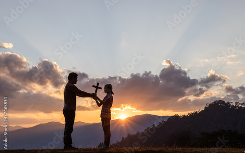 Canvas Print Silhouette Christian father holding Christian cross teaching his daughter to pray outside on mountain sunset background