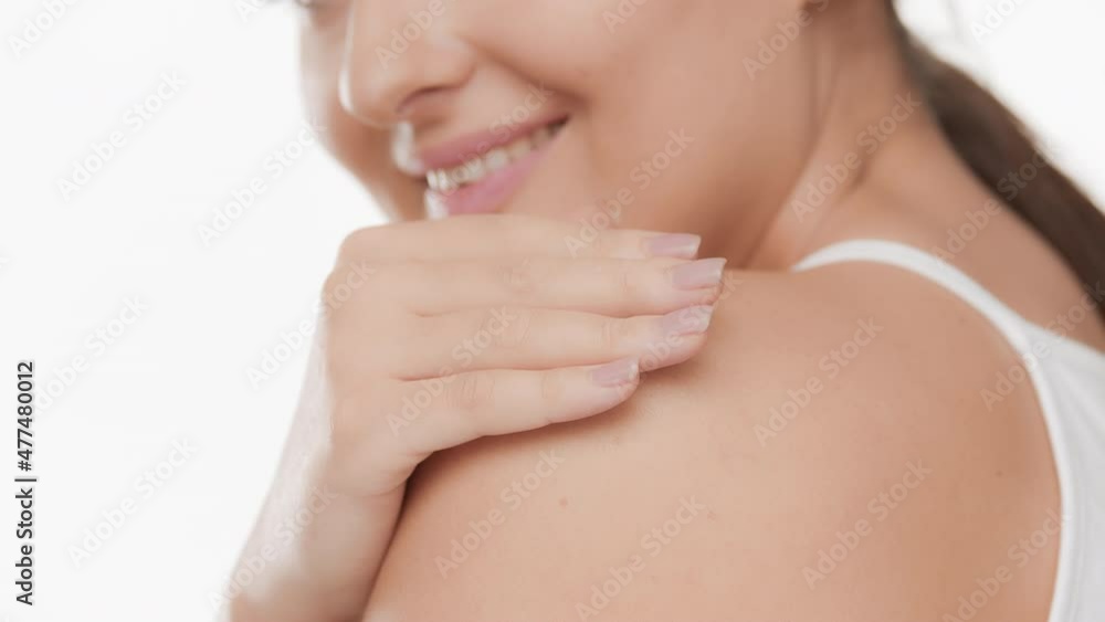 Good-looking white-skinned young brunette plus size woman with long ponytail in white top touches her shoulder smiling wide for the camera on white background | Body care concept Good-looking white-skinned young brunette plus size woman with long ponytail in white top touches her shoulder smiling wide for the camera on white background | Body care concept