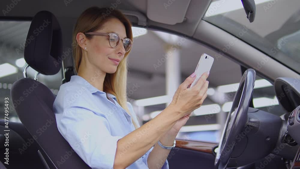 Beautiful self-confident blond woman in stylish shirt sitting on driver seat of her respectable car and scrolling web-pages on smartphone, doing shopping online, checking bank account Beautiful self-confident blond woman in stylish shirt sitting on driver seat of her respectable car and scrolling web-pages on smartphone, doing shopping online, checking bank account
