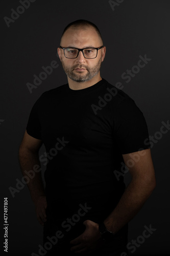 A young adult man in black-rimmed glasses and a black T-shirt poses against a dark background, looking intently at the camera © dimetradim