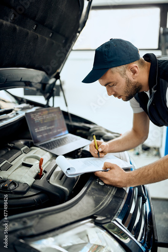 Young repairman takes notes while running diagnostic of car engine in workshop.