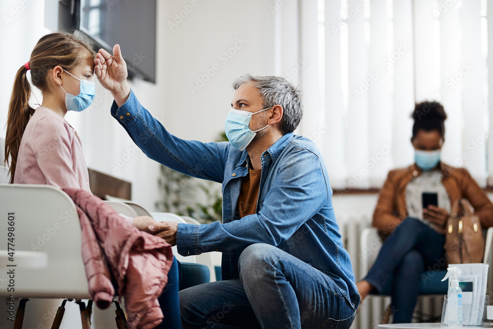 Worried father checks daughter's forehead for fever in waiting room at doctor's office during coronavirus pandemic. Worried father checks daughter's forehead for fever in waiting room at doctor's office during coronavirus pandemic.