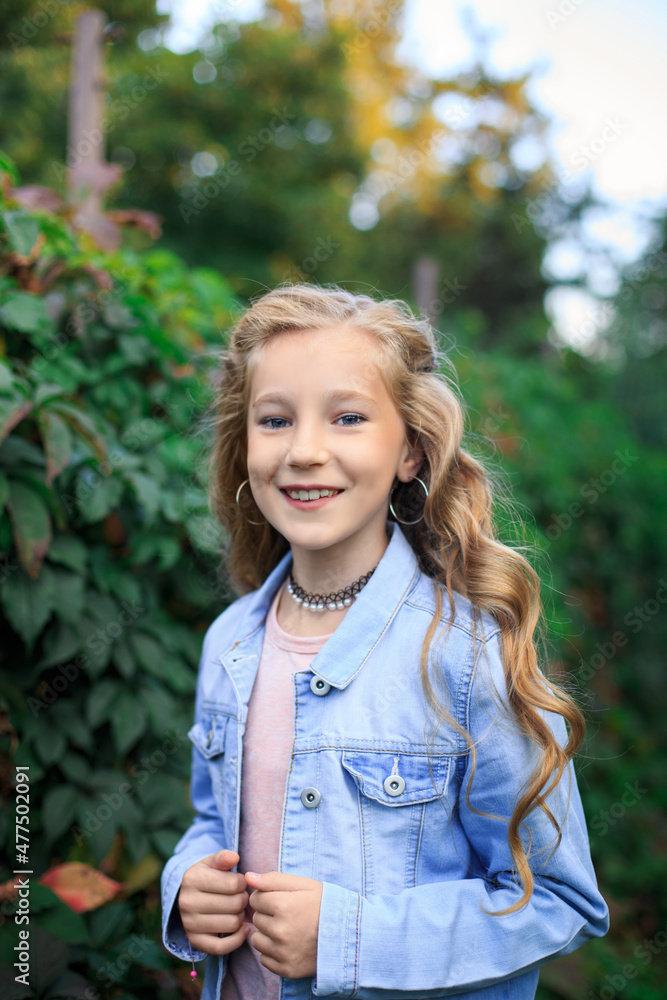 Portrait of a joyful blond-haired, blue-eyed girl against a background of lush green bushes. The girl is wearing a blue denim jacket Portrait of a joyful blond-haired, blue-eyed girl against a background of lush green bushes. The girl is wearing a blue denim jacket