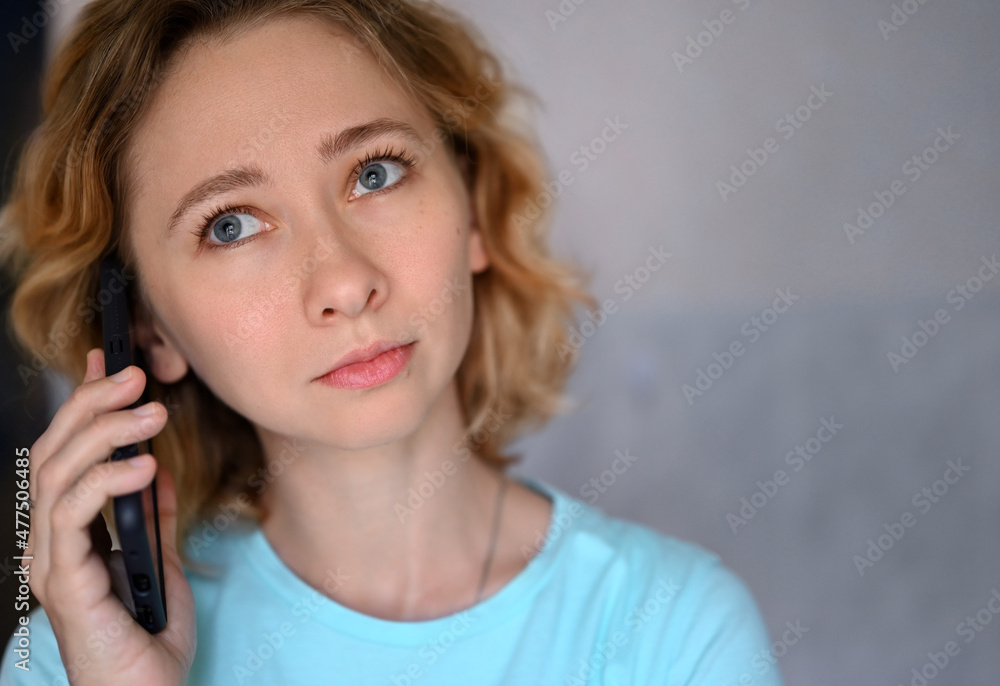 Close-up portrait of young caucasian woman talking on cell phone at home.The blue-eyed blonde looks up thoughtfully. She is wearing a blue T-shirt. Close-up portrait of young caucasian woman talking on cell phone at home.The blue-eyed blonde looks up thoughtfully. She is wearing a blue T-shirt.