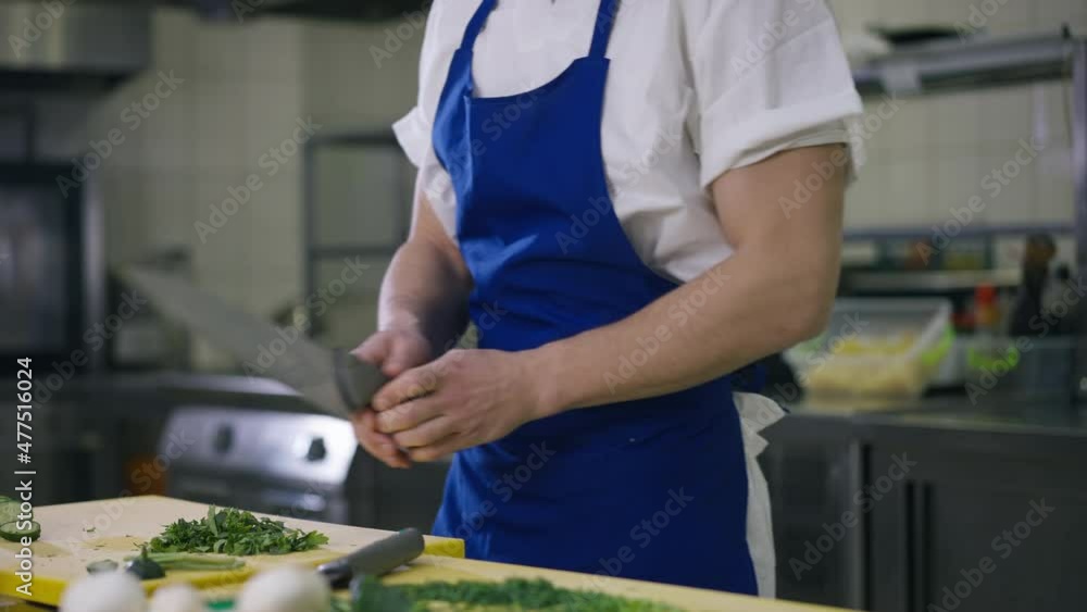 Unrecognizable male cook cleaning kitchen knife rubbing utensil on apron and cutting greenery. Young Caucasian man cooking delicious salad preparing ingredients in restaurant indoors Unrecognizable male cook cleaning kitchen knife rubbing utensil on apron and cutting greenery. Young Caucasian man cooking delicious salad preparing ingredients in restaurant indoors
