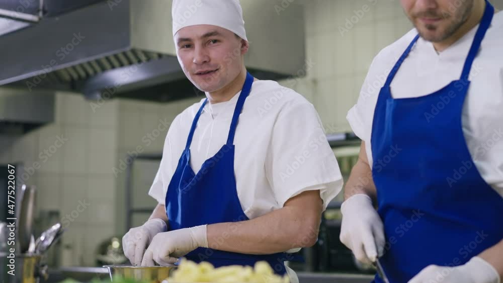 Positive man talking mixing salad in bowl and smiling looking at camera standing with unrecognizable colleague in kitchen. Portrait of confident cook in uniform indoors at workplace with coworker Positive man talking mixing salad in bowl and smiling looking at camera standing with unrecognizable colleague in kitchen. Portrait of confident cook in uniform indoors at workplace with coworker
