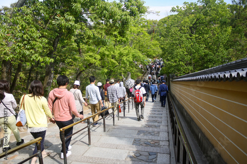 Crowd of people walking down pathway inside zen garden of ancient temple in Kyoto, Japan with trees foliage and blue sky background.