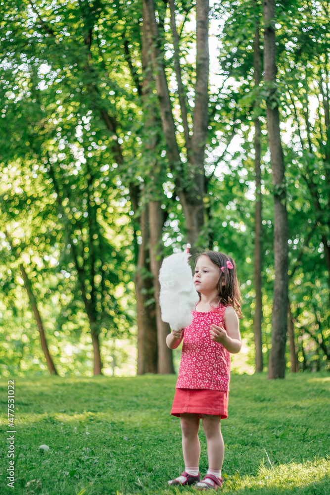 Little cute girl 3-4 eating cotton candy in sunny park among tall trees on green grass. Vertical