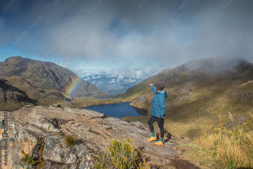 horizontal shot of a hiker looking towards landscape full of paramo vegetation with rocks and a beautiful lake with a blue sky and rainbow in Chirripo National Park in Costa Rica