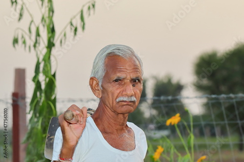 An senior male Indian farmer standing in the field at sunset with a shovel on his shoulder and wore a white vest and looking at camera