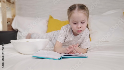 little child eats reading book while lying bed, happy family, kid daughter doing homework and eating potato chips, doing homework elementary school, first grader loves literature, children school life © Валерий Зотьев