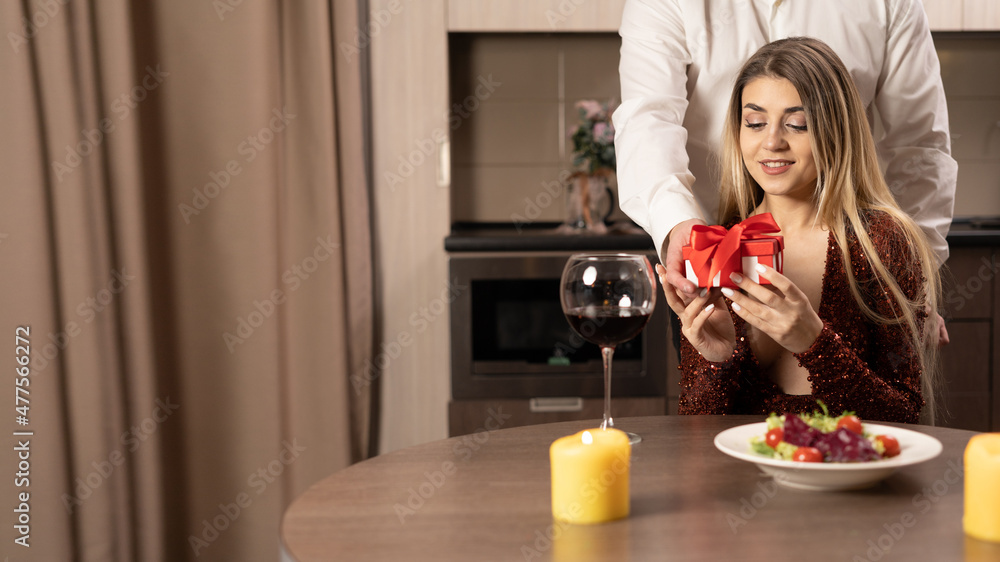 Happy young woman receiving a gift box from a loving man. A loving couple happily celebrates Valentine's Day, enjoying a romantic dinner, holding the present moment, sitting at the home table. Happy young woman receiving a gift box from a loving man. A loving couple happily celebrates Valentine's Day, enjoying a romantic dinner, holding the present moment, sitting at the home table.