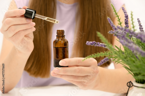 Unrecognizable caucasian woman with long brown hair sitting at table holding glass bottle, applying natural serum,oil on hair or skin. Bio cosmetic,beauty procedures at home concept,close-up